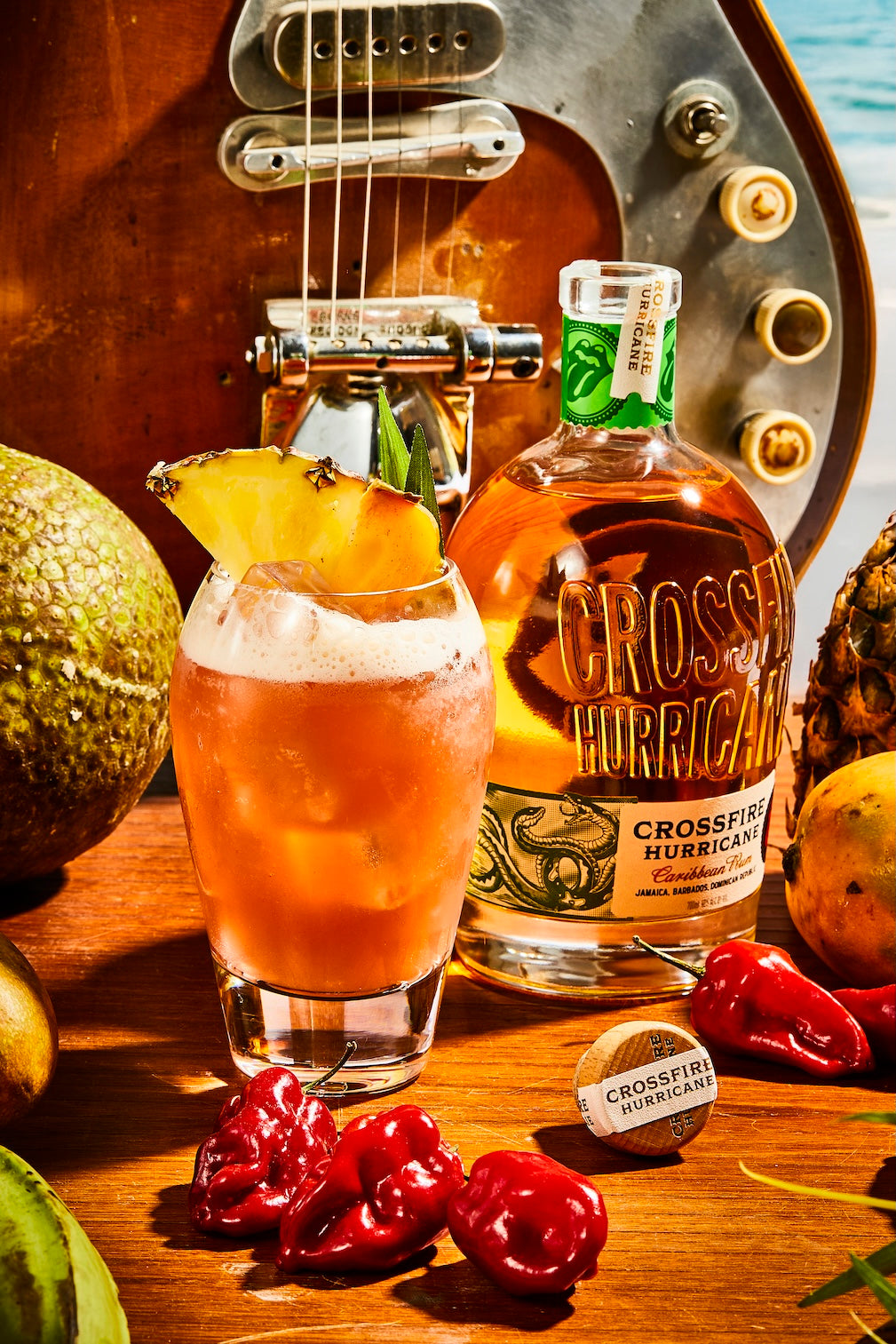 Crossfire Hurrican Rum sitting next to a cocktail on a table full of fruit with a guitar in the background.